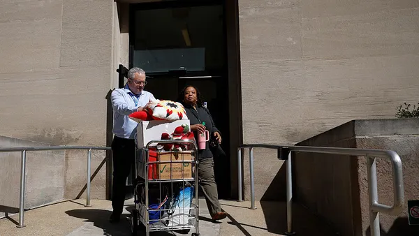 Two people wearing work attire exit a building with a cart of work items.