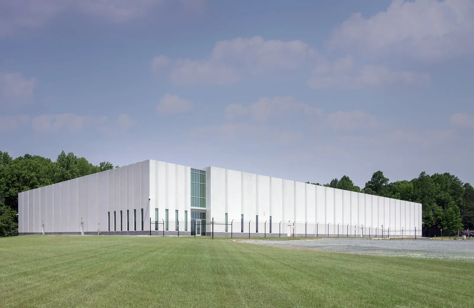 A white building with green grass and a fence in front of it, and green trees behind it.