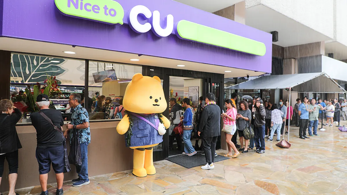 A line of people wait to get into a store. A sign over the door says "Nice to CU" in purple and while lettering and a bear mascot stands outside near the door.