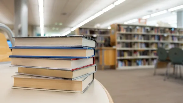 A stack of books sits on a table in the foreground. In the background are rows of shelves of books in a room