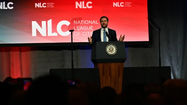 Man is a suit at a podium in front of a sign that reads "NLC - National League of Cities"