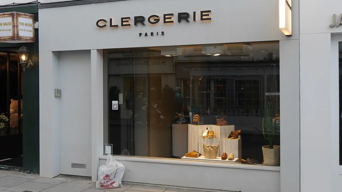 Closed Clergerie shoe store on a cobblestone street with darkened windows and a few shoes on stands on display. A plastic grocery bag sits in front of the store.