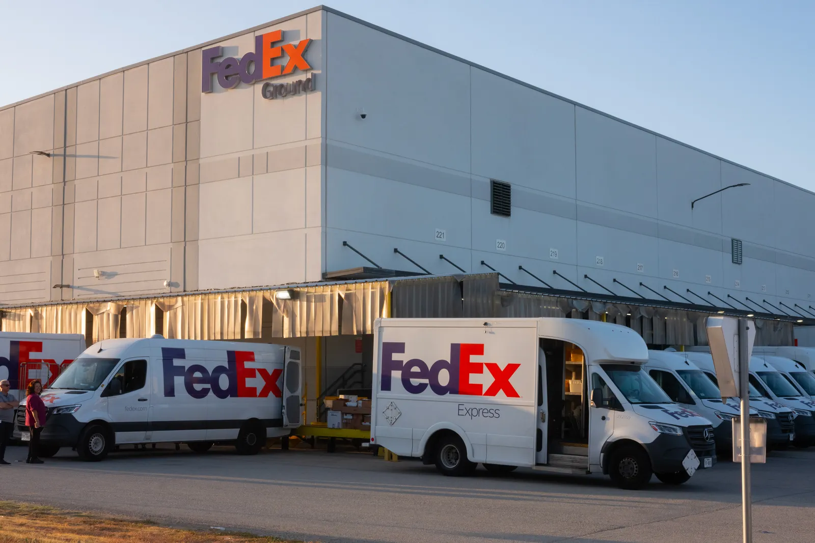 A wide shot of a large FedEx warehouse building featuring signs that read "FedEx Ground" and "FedEx Express" during a bright orange sunrise. Several white delivery vans with the purple and orange "FedEx" logo are parked at the loading docks.
