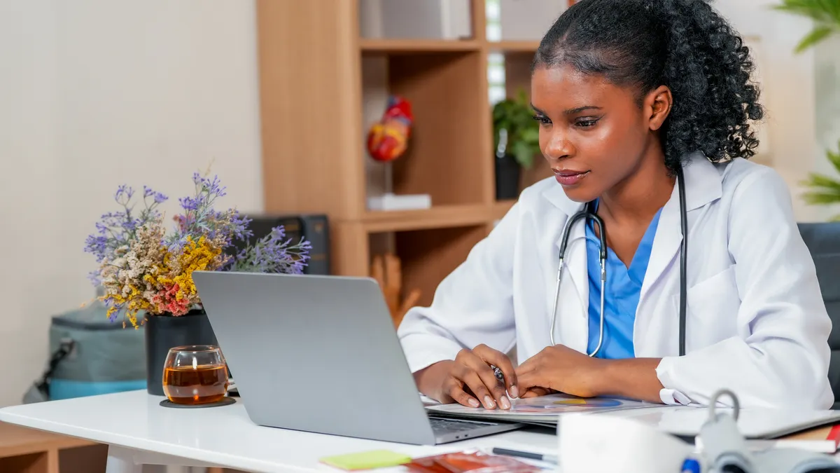 A focused doctor in a white coat, with a stethoscope, works on a laptop at a desk.