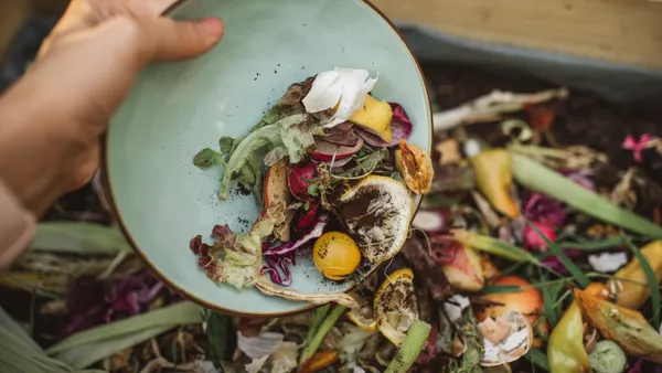 A hand brushes organic materials like vegetable scraps off a plate and into a bin