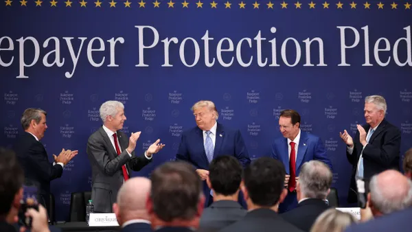 US President Donald Trump stands on stage while people around him applaud. The word "protection" can be seen behind him.