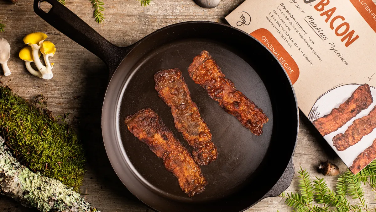 Overhead view of three strips of fried MyBacon in a cast iron skillet on a wooden surface. Moss, branches and mushrooms are arranged around the pan, and the package is in the top right corner.