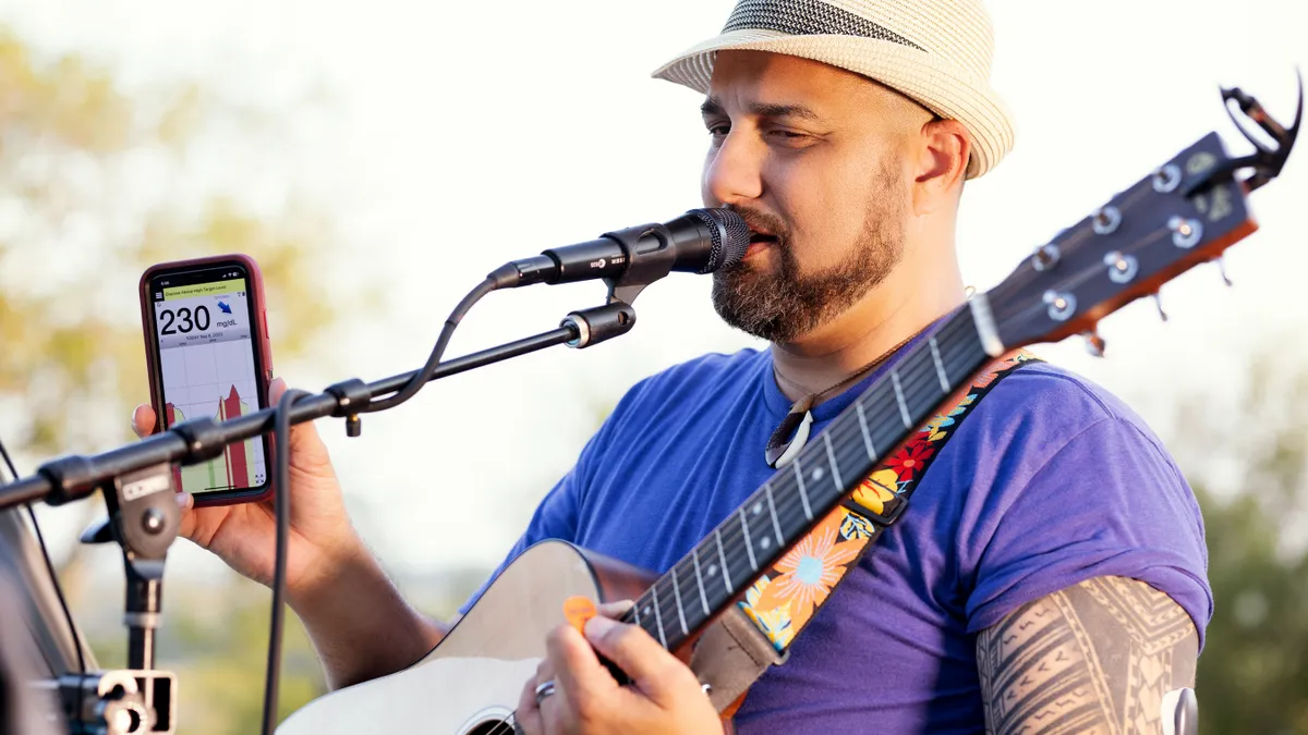 A person playing a guitar in front of a microphone holds up a phone with a blood glucose reading on it.