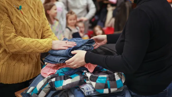 A woman volunteer helps another woman sort clothes on a table in a charity setting. Several children are in the background.