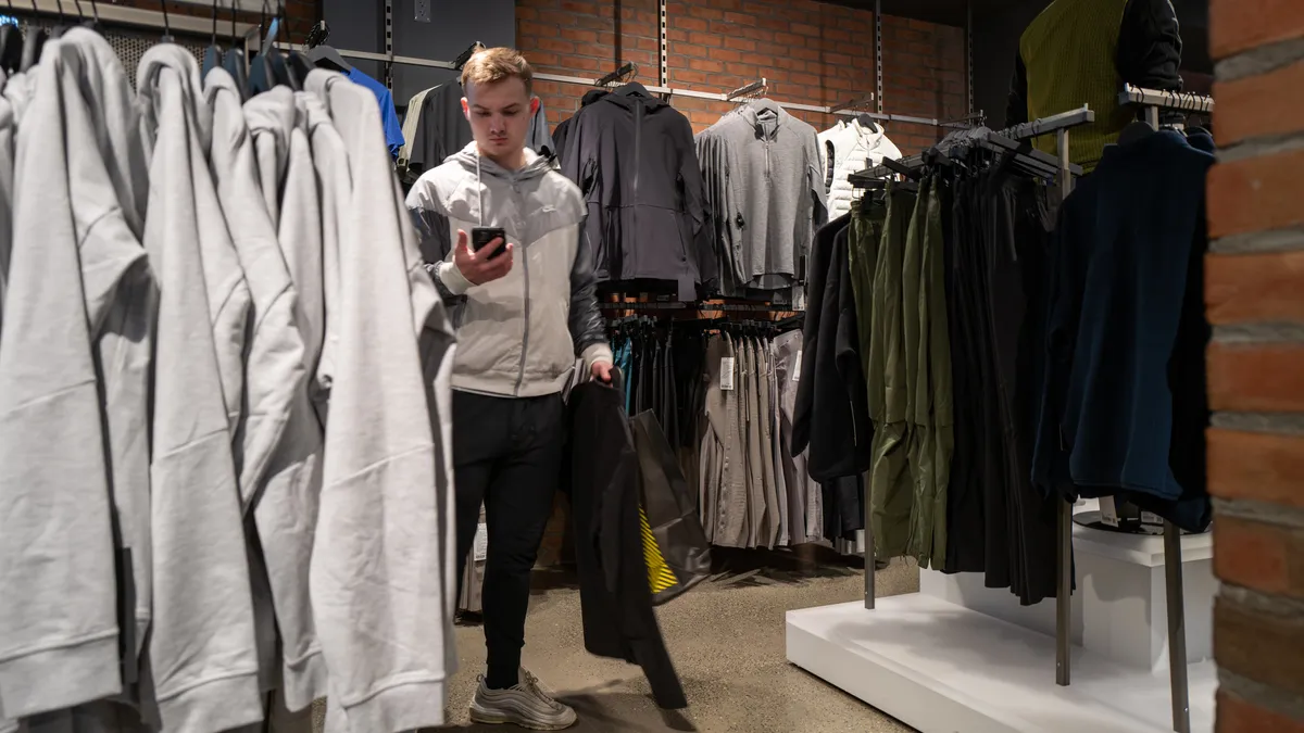 A person is standing in the men's section of a clothing store, holding clothing items.