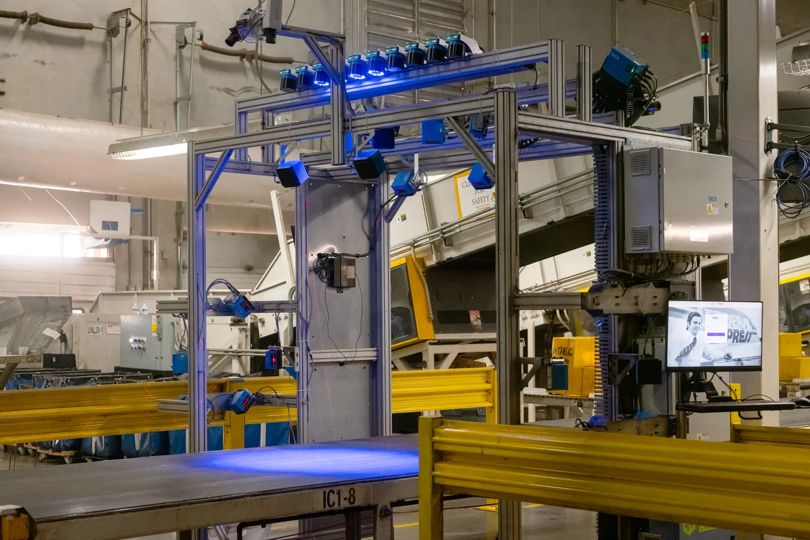 A large, metal digital scanning structure with multiple blue-lit cameras is positioned over a conveyor belt labeled "IC1-8" inside a warehouse. Yellow safety railings are in the foreground, and a computer monitor to the right displays a FedEx screen.