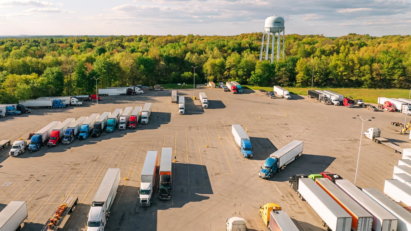 Dozens of tractor-trailers parked at a rest stop with a water tower and green trees in the background.