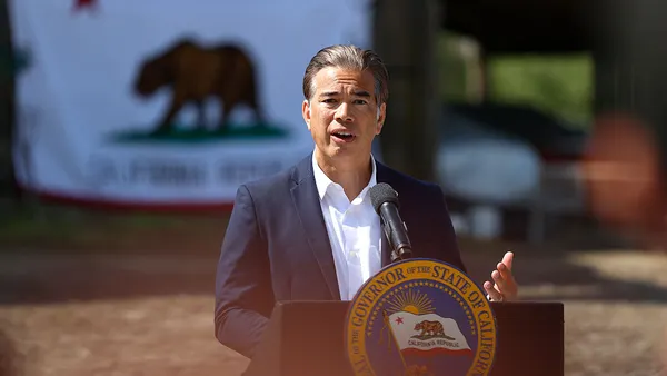 A person stands behind a podium that has a microphone. A state flag of California is seen blurred in the background