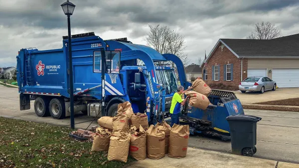 A residential neighborhood scene featuring a blue Republic Services garbage truck collecting compostable yard waste. A worker in a bright yellow vest loads large brown paper leaf bags into the truck. A row of suburban houses and overcast skies form the backdrop.