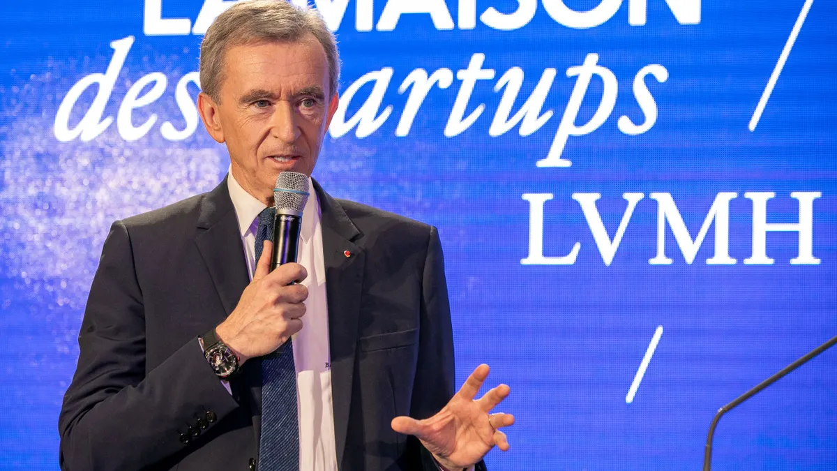 Bernard Arnault wears a suit and holds a microphone while standing against a blue background with white lettering.