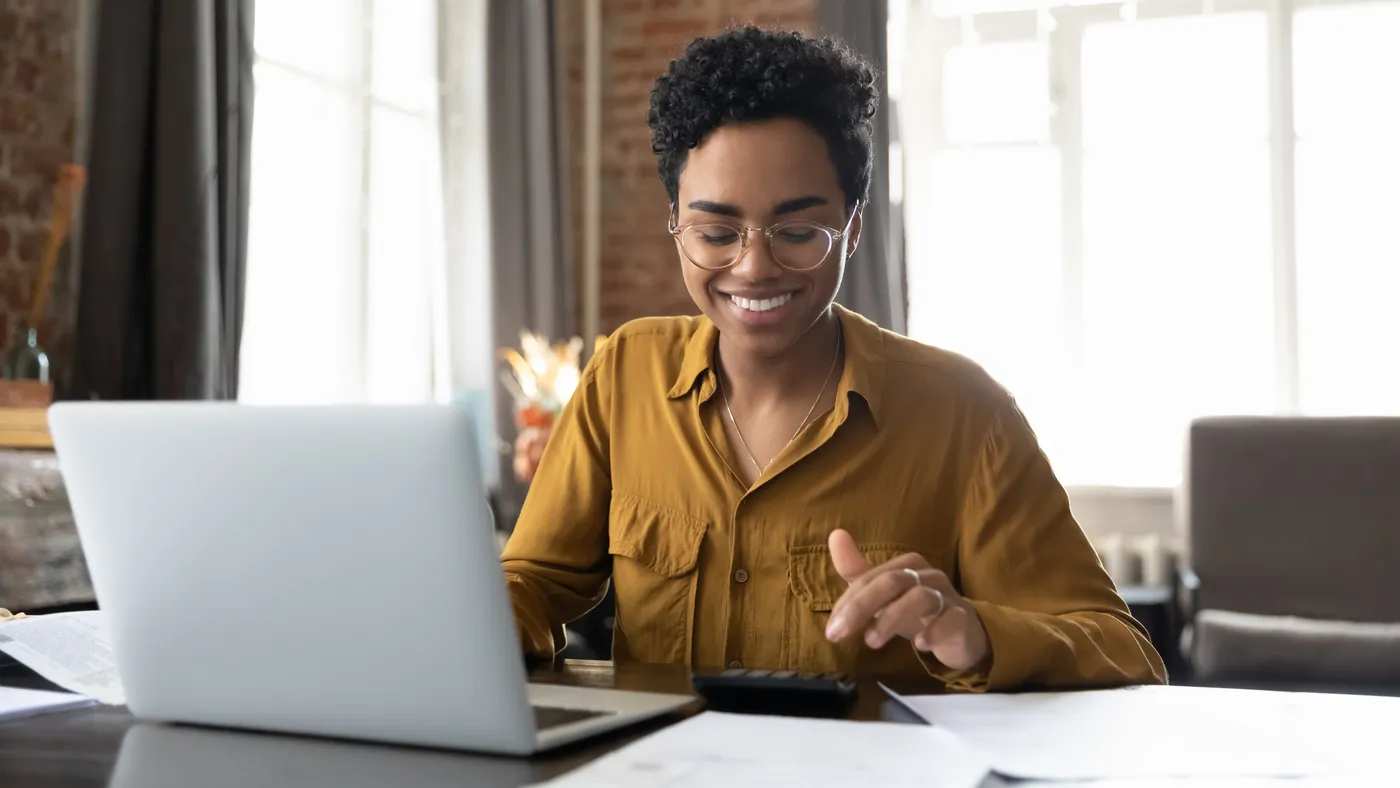 A person sits in front of a laptop and smiles while using a calculator.