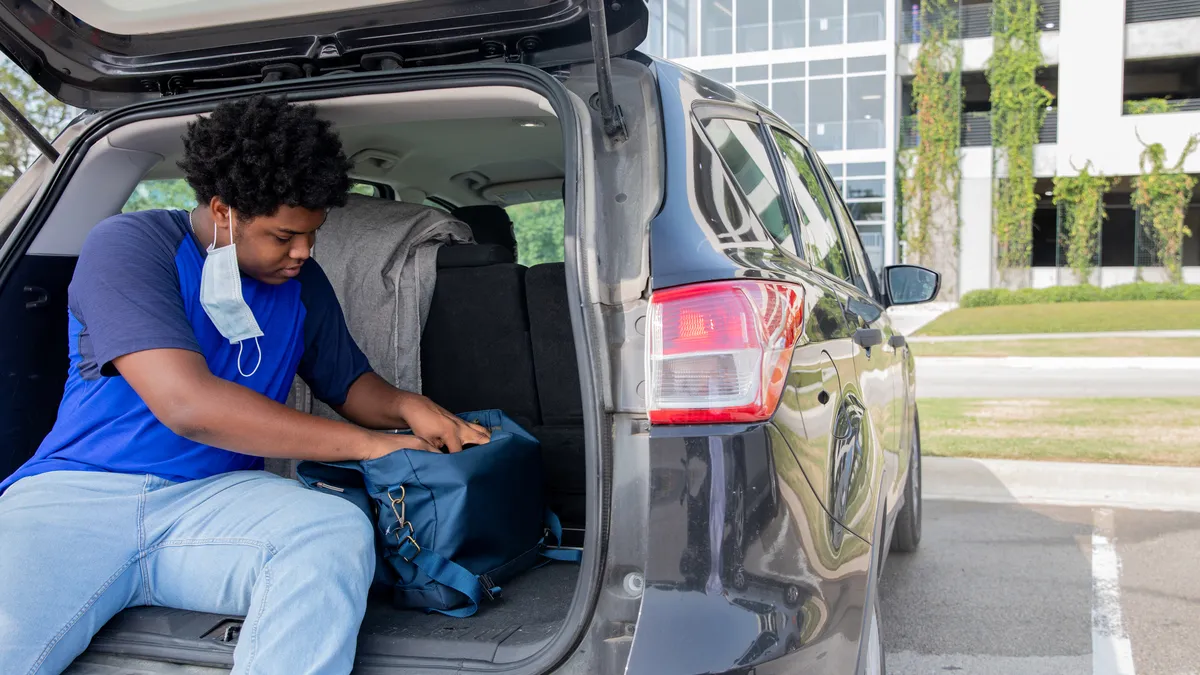 A boy with a mask hanging from his year reaches for his backpack in the trunk of a car