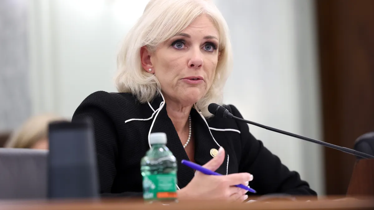 A blonde woman in a black suit speaking into a mic with a purple pen in her right hand.