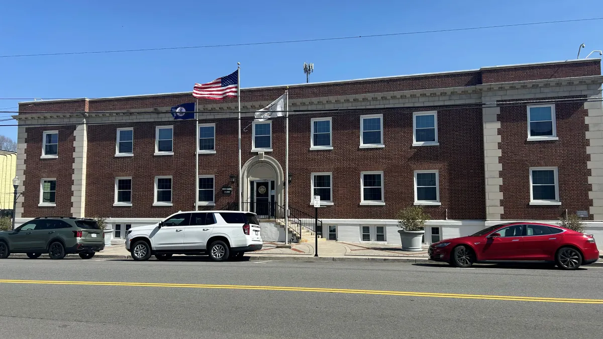 A two-story red brick building on a multilane street with a U.S. and other flag visible and cars parked in front of it