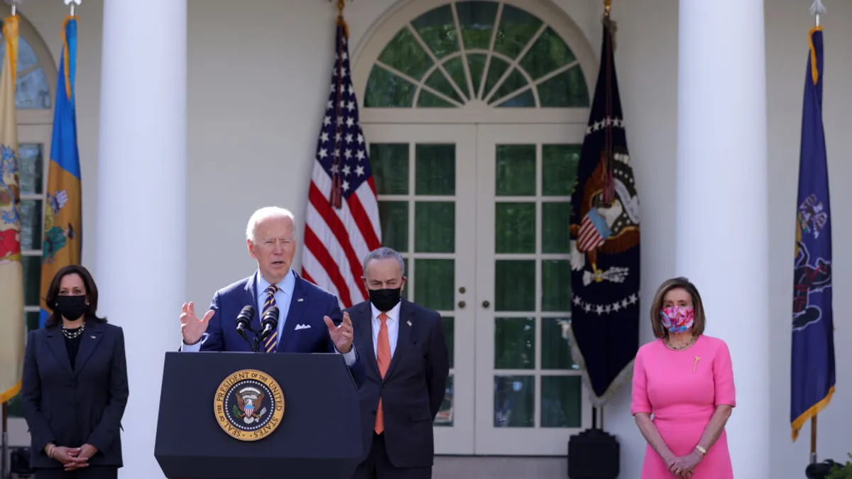 A person in a suit stands at a podium with a microphone. Behind him are flags and three people wearing face masks.