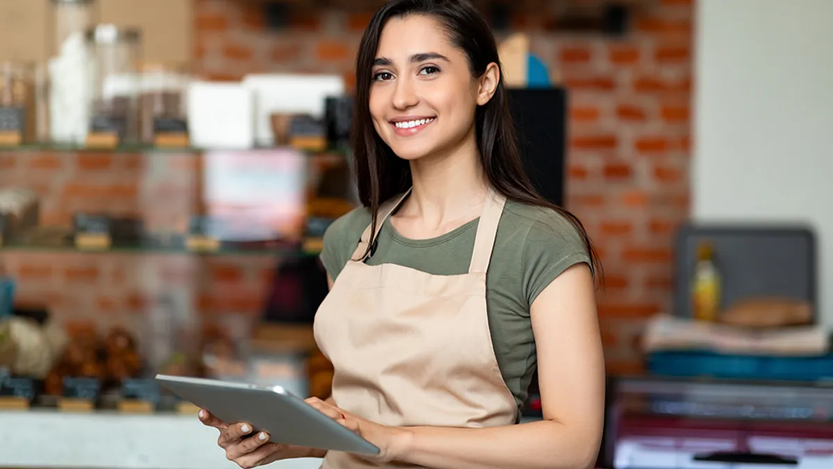 Opening small business. Happy arab woman in apron near bar counter holding digital tablet and looking at camera.