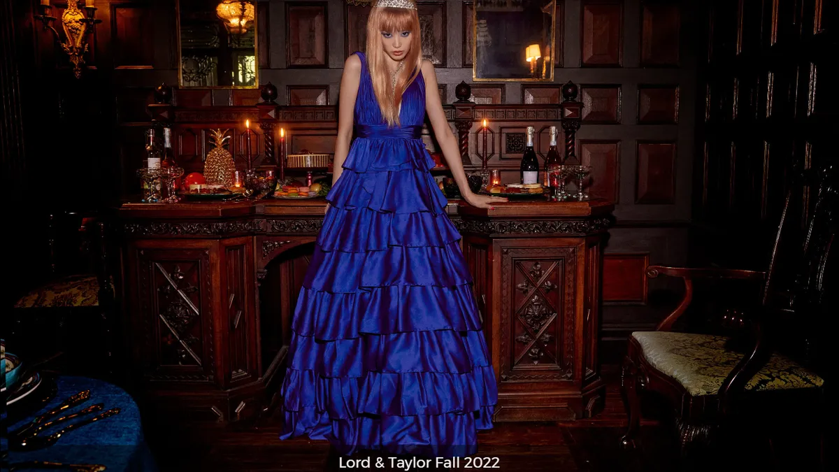 A model in a blue dress and tiara leans against a desk