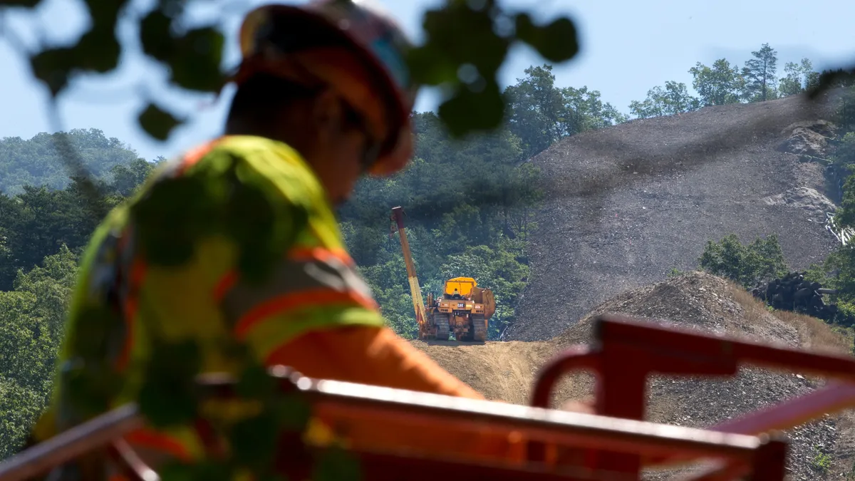 A person in a reflective vest is in the foreground, while a large piece of yellow construction equipment moves along a dirt swath through a mountain in the background.