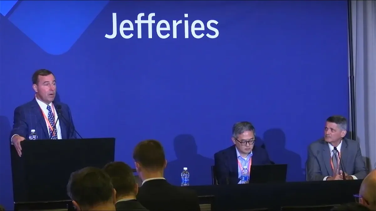 A person stands at a podium while speaking, next to two people sitting at a table, in front of a blue background with the word Jefferies in white.