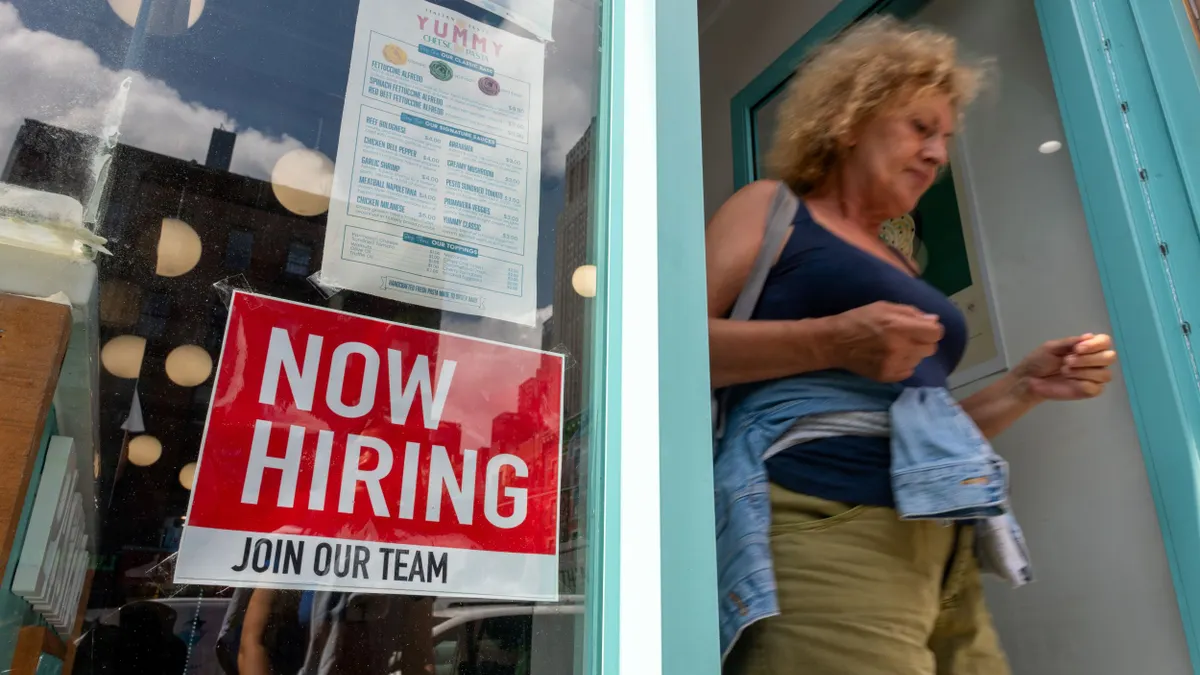 A 'now hiring' sign is displayed in a business window as a person exits through a door.