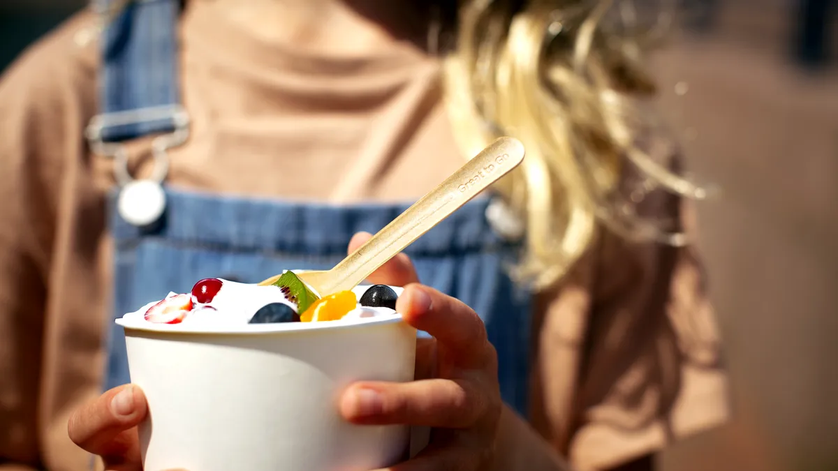 Hands hold a white paper cup filled with yogurt and fruit