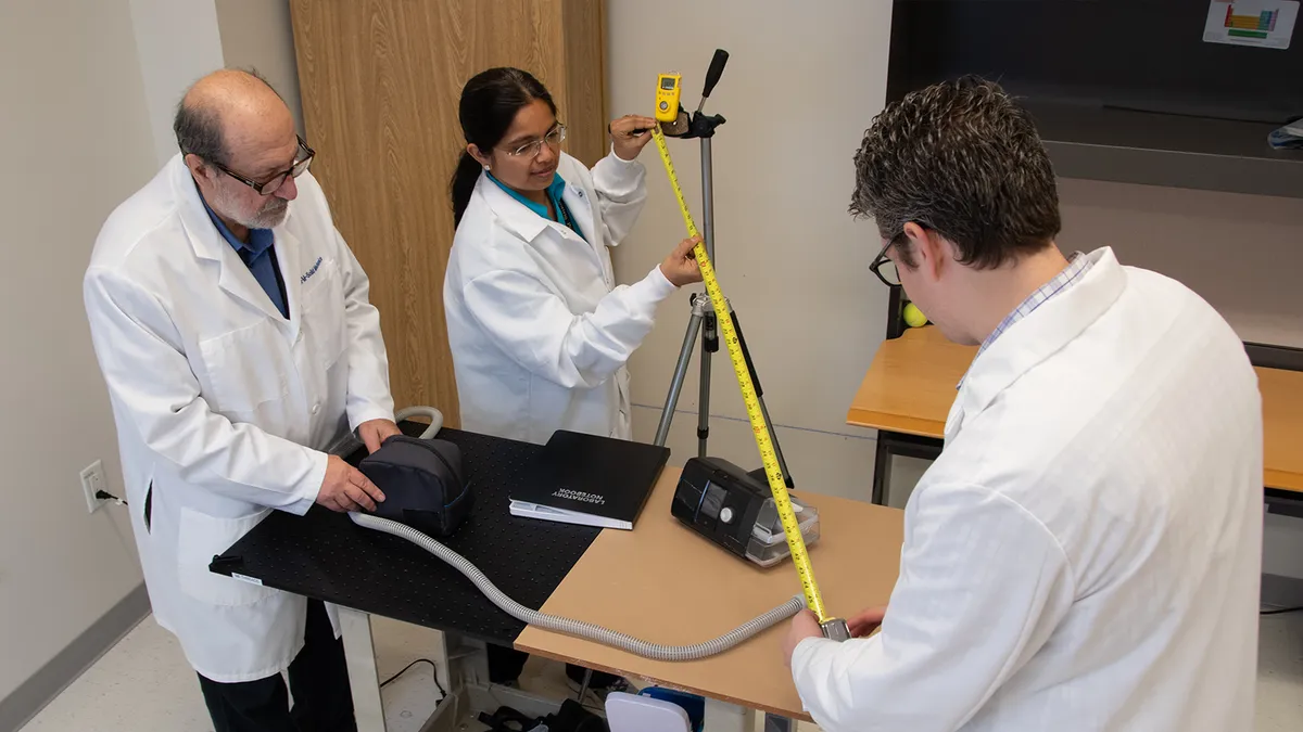 Three people stand aroud a table wearing white coats. They hold a tubed device and a measuring tape.