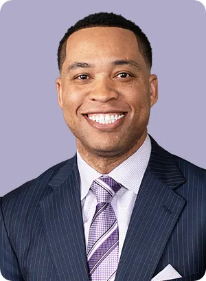 A person wearing a dark suit and a purple tie smiles.