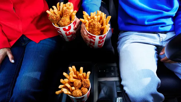 A photograph of the inside of a car where two people are sitting. In the center there is small cups of chicken and fries.