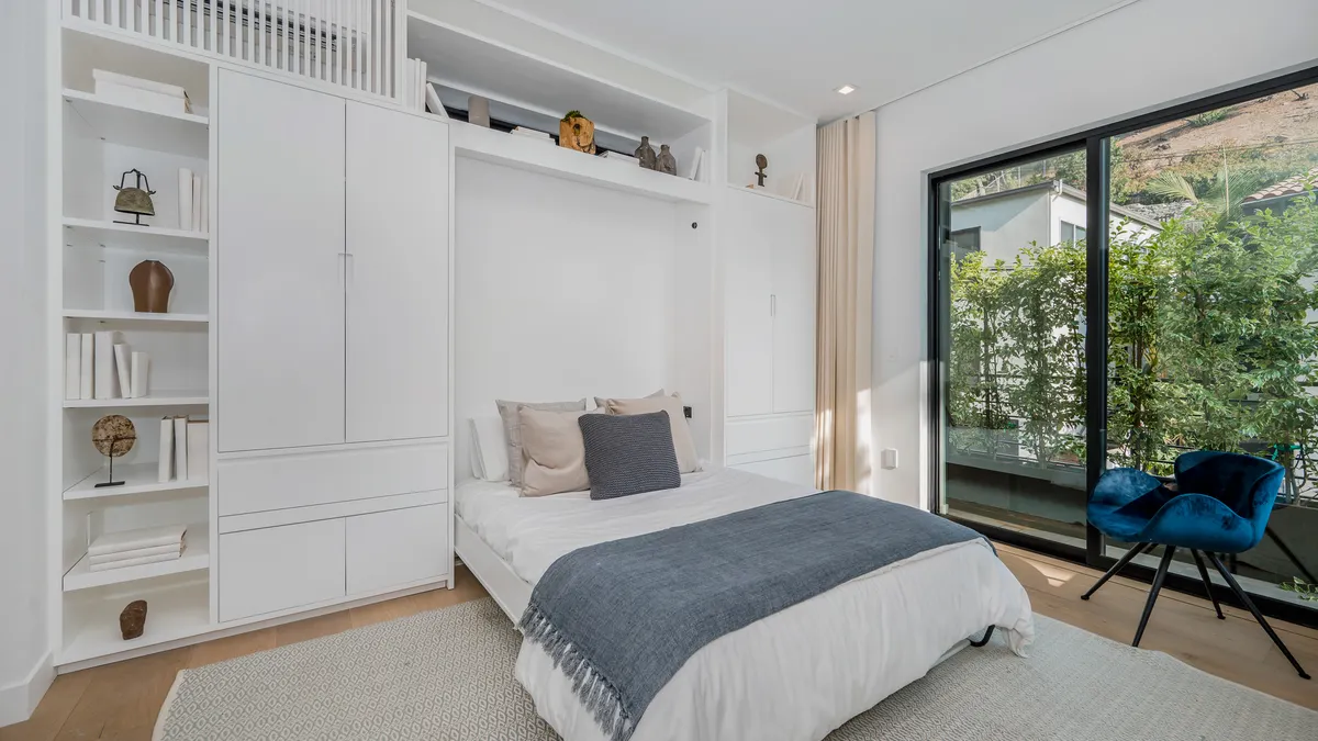A bedroom with a wall bed, chair and shelves above the bed.