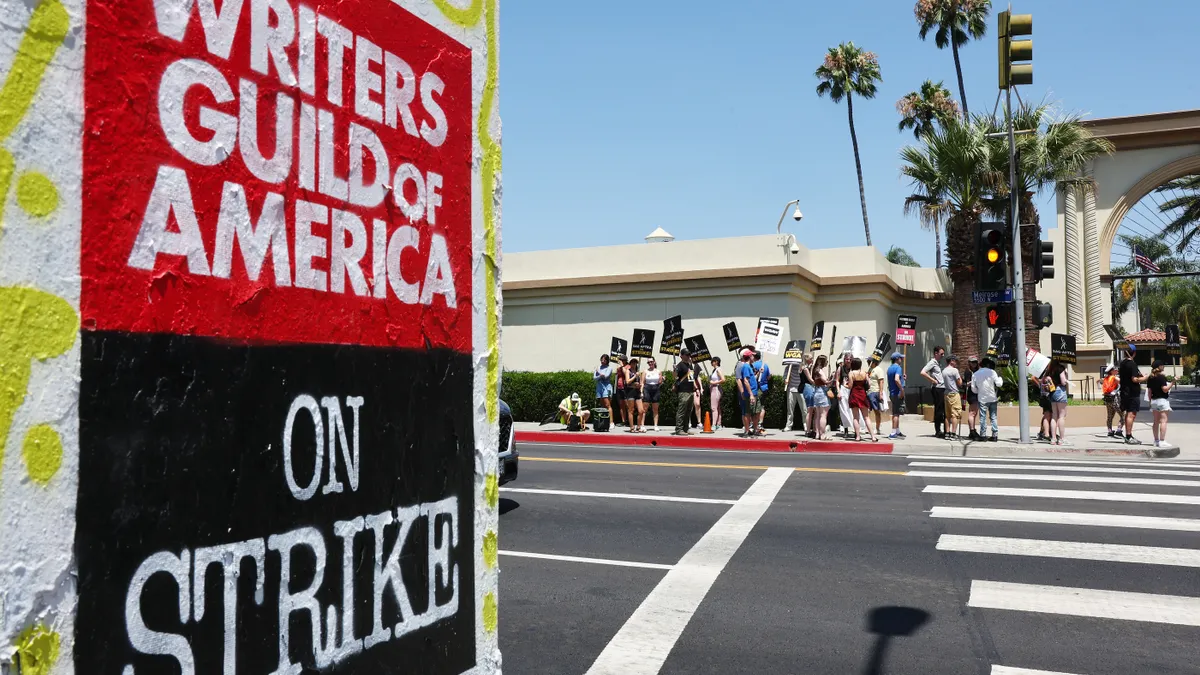 Graffiti says "WRITERS GUILD OF AMERICA ON STRIKE"; picketers across the street visible with blue skies and palm trees