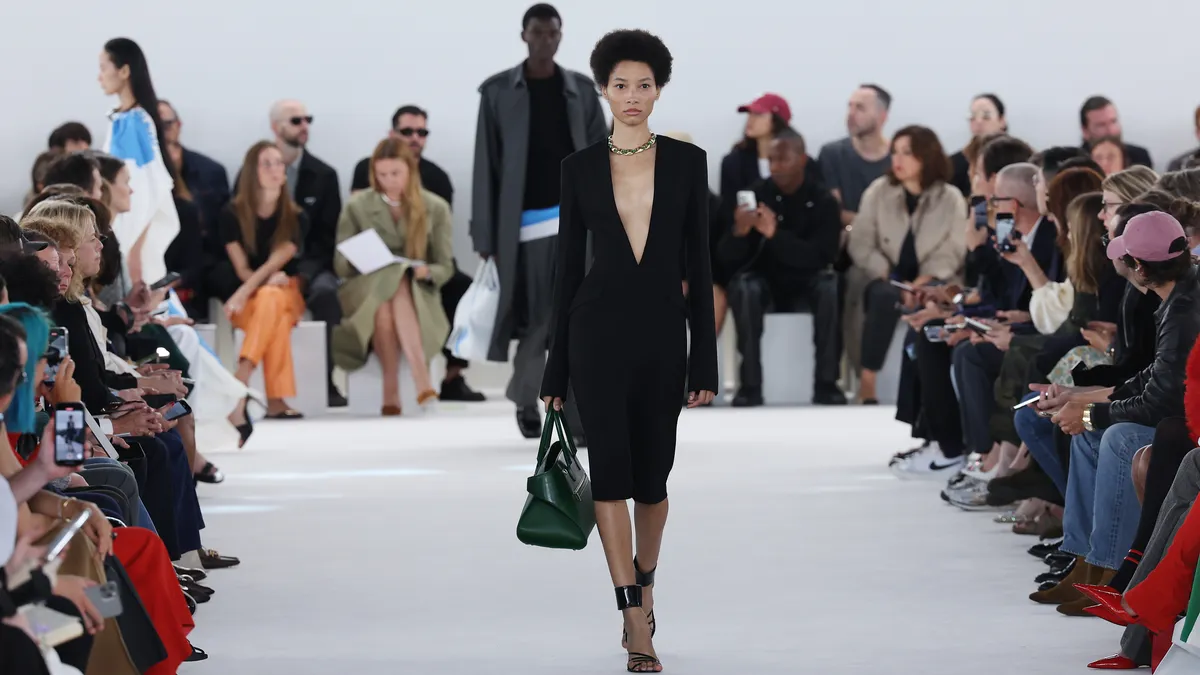 A model wears a black dress and walks on a runway as a crowd watches from the sidelines.