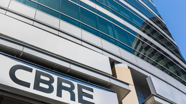 Upward view of a modern glass and steel office building with the CBRE logo prominently displayed on a sign at the entrance. The structure features reflective blue-tinted windows, and another curved building is reflected in the glass façade.