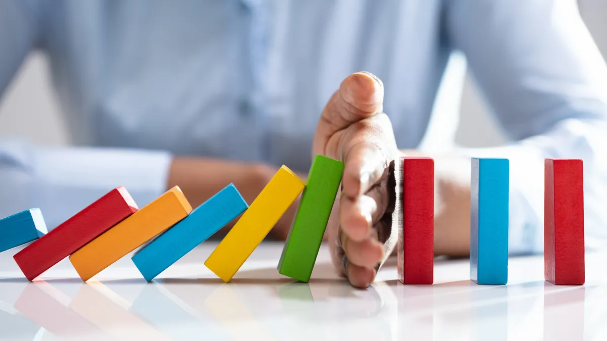 Close-up Of A Businesswoman Hand Stopping Colorful Dominoes From Falling On Office Desk