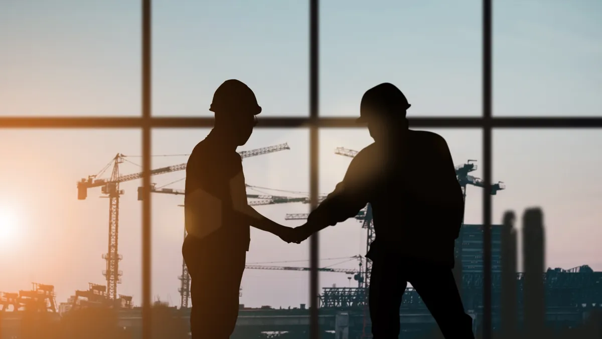 Stock image of two construction workers silhouetted against a window with construction cranes in the background.