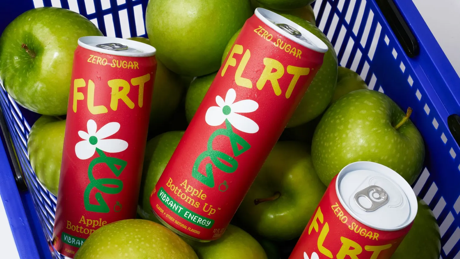 Three cans of Flrt are in a shopping basket with green apples.