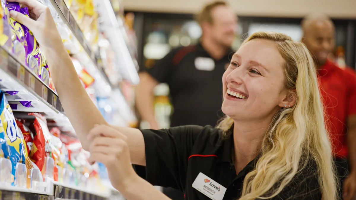 A photo of a person inside a store, arranging items on a shelf. The person is wearing a nametag that reads "Love's" and "Meredith."