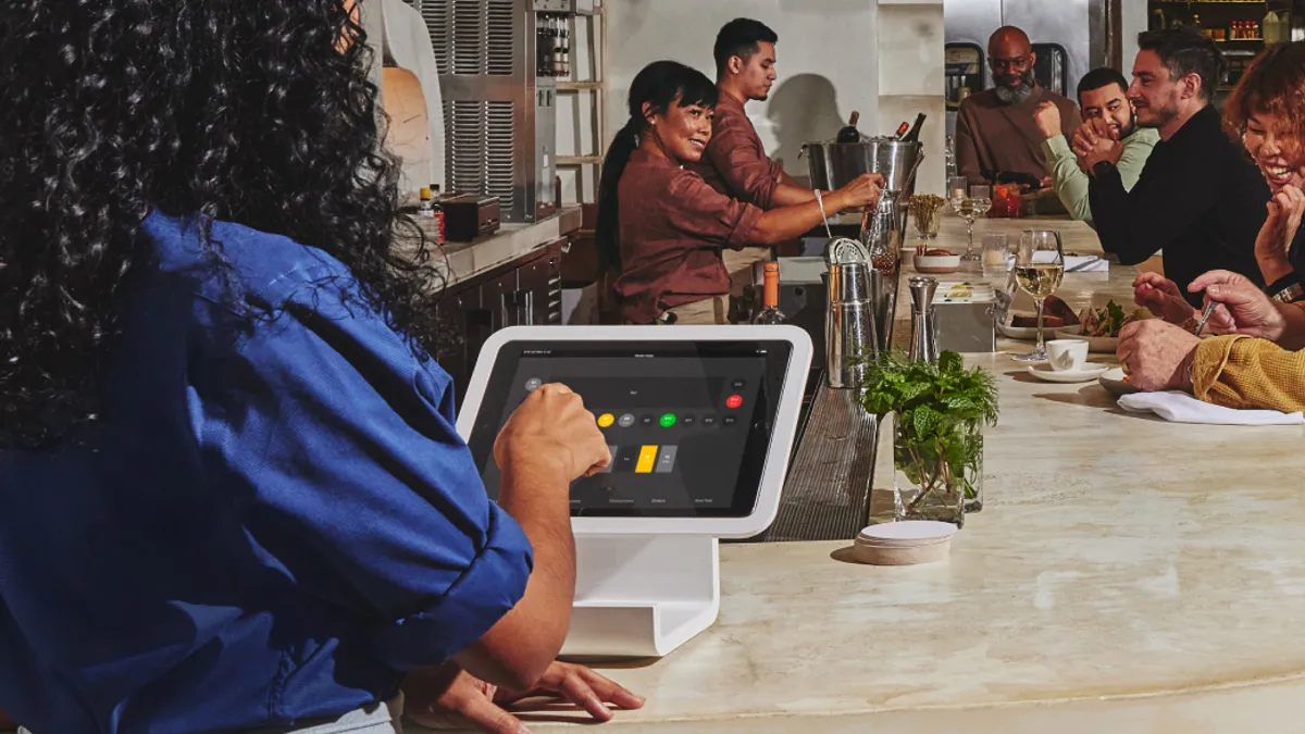 A restaurant worker uses a tablet for payment operations while restaurant customers mingle in the background.