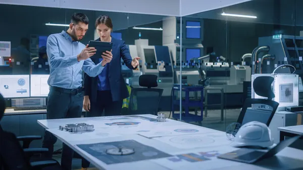 Factory Office Meeting Room: Male Chief Engineer Holds Digital Tablet Computer, Shows Screen to Female Project Manager, They Talk, Find Optimization Solution.