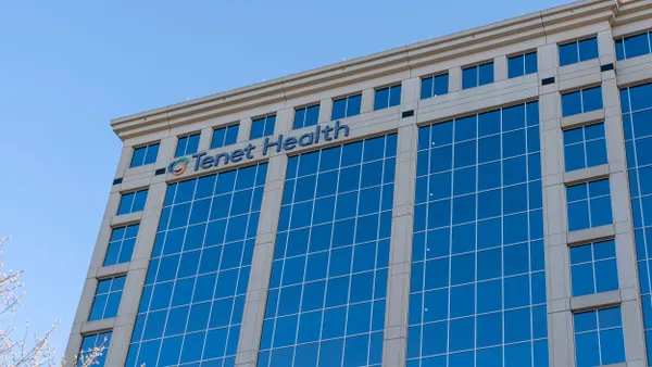 A tall, modern office building with a facade made up mostly of reflective blue glass windows. The “Tenet Health” logo is displayed near the top corner of the structure. The sky is clear and blue, enhancing the building's sharp, corporate appearance.
