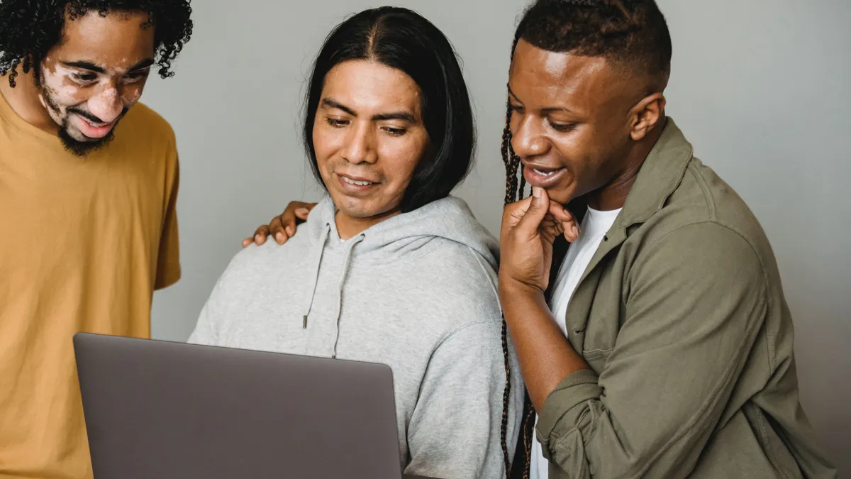 Three brown people stand side-by-side looking at a laptop