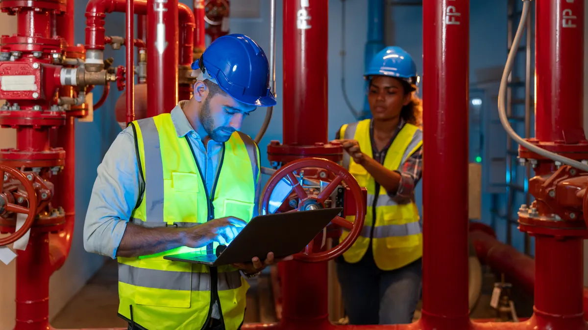 A building engineer uses his laptop to inspect the pipeline and valve of service water supply in a fire riser room, while a technician in the background tests a fire suppression system.