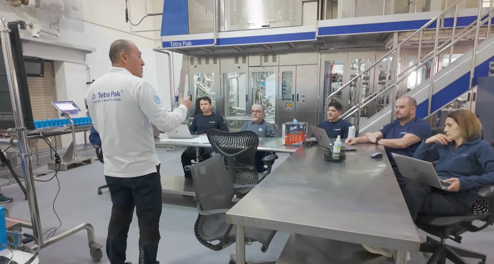 One person stands and speaks to five people seated at a metal table in an industrial setting with a Tetra Pak logo on the wall.