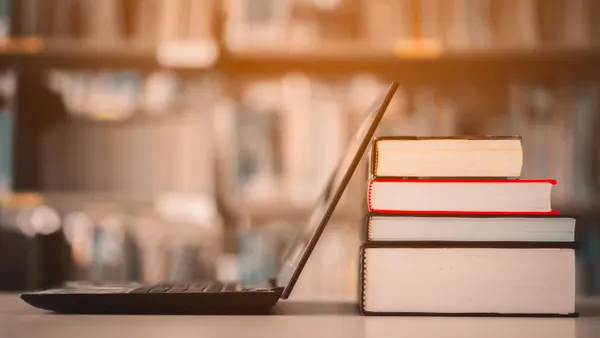 An open laptop screen rests on a stack of printed books, which are on a table. In the background seems to be a bookshelf.