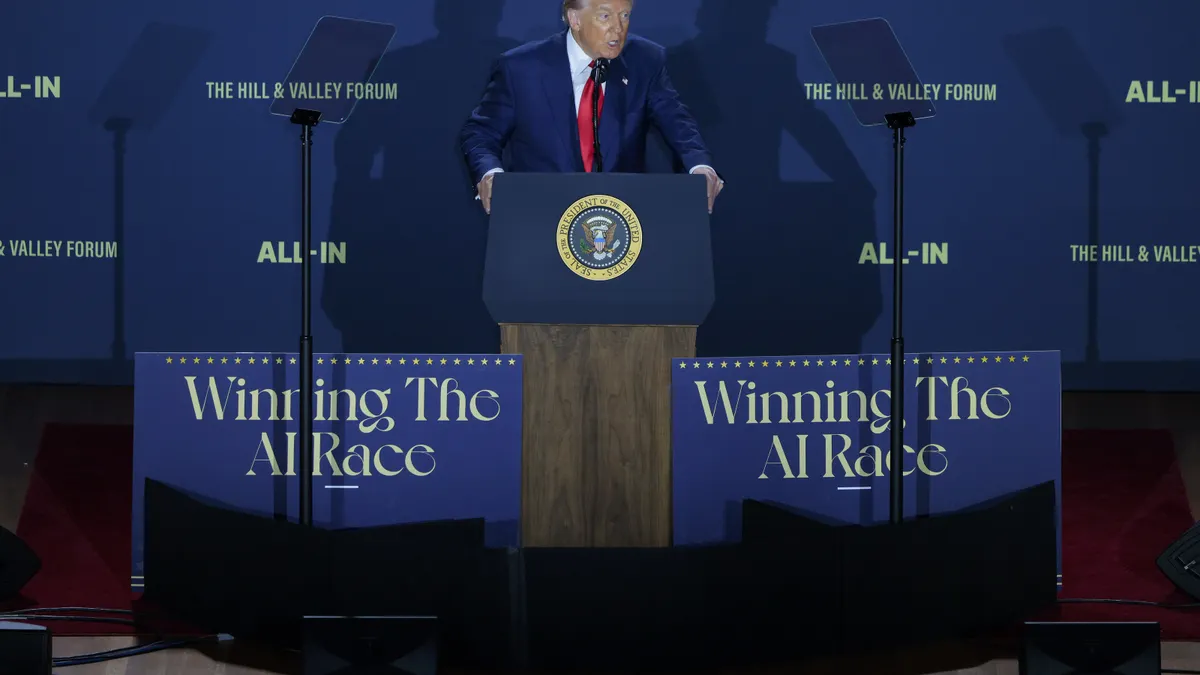 President Donald Trump stands at a lectern on a podium in front of a blue backdrop. Signs in front of him say "Winning the AI race" and the backdrop behind him says "The Hill & Valley Forum" and "All-In."