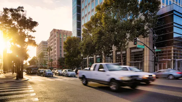 Vehicles and a bus on a multilane street lined with trees and tall buildings. The sun flares on the left behind the trees.